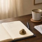 A leather-bound notebook open to a page headed Essentials, with a brass paperweight, navy fountain pen, and brass-rimmed coffee mug on a wooden desk in soft morning light