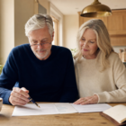 White couple in their early 60s reviewing a pension election letter and a one-page retirement income summary at a sunlit kitchen table, brass pendant lamp and brass-rimmed cream mug nearby — bucket planning with a pension featured image