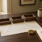 Editorial still-life: three small empty wooden trays lined up left to right at slightly increasing heights, a folded cream paper with a navy-ink architectural foundation diagram, brass building cube blocks, paperweight, leather notebook with navy fountain pen, brass-rimmed cream mug on a warm walnut desk in soft morning light.